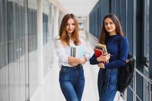 dos joven mujer con libro chateando mientras en pie en Universidad corredor. Universidad estudiantes en corredor después el conferencia. foto