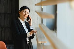 alegre joven negocio mujer en pie en moderno oficina hablando en su móvil teléfono. hermosa hembra modelo utilizando célula teléfono. foto