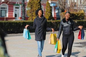 dos afro americano mujer amigos en el ciudad en un compras viaje que lleva vistoso compras bolsas. foto