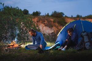 noche cámping en el montañas. contento Pareja viajeros sentado juntos junto a hoguera y brillante turista carpa. en antecedentes grande roca, bosque y noche cielo. foto