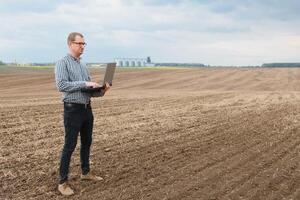 cosecha concepto. granjero en un campo con un ordenador portátil en un antecedentes de un agrícola silos para almacenamiento y el secado de granos, trigo foto