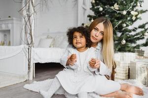 alegre Navidad y contento Días festivos alegre mamá y su linda hija a Navidad árbol. padre y pequeño niño teniendo divertido cerca Navidad árbol adentro. amoroso familia con regalos en habitación. foto