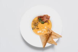 Pumpkin cream soup. White ceramic bowl with fresh pumpkin, dry pumpkin seeds, smoked bacon on a white background photo