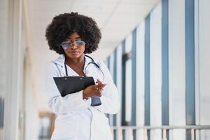 Closeup headshot portrait of friendly, smiling confident female healthcare professional with lab coat, arms crossed holding glasses. Isolated hospital clinic background. Time for an office visit photo