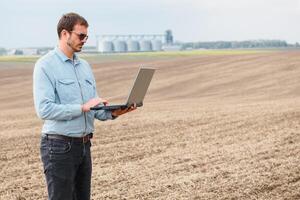 cosecha concepto. granjero en un campo con un ordenador portátil en un antecedentes de un agrícola silos para almacenamiento y el secado de granos, trigo foto