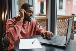 afro americano negociaciones vídeo charla en computadora se sienta en café con taza de café. negro hombre llamadas en ordenador portátil en vídeo enlace y habla él usa en camisa y traje chaqueta. vídeo enlace en línea conexión foto