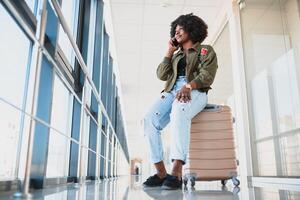 Portrait of happy young african american woman sitting on suitcase and talking with mobile phone at the station photo