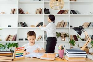 Young female tutor helping little elementary school boy with homework during individual lesson at home photo