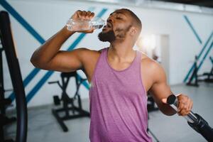 young african american sportsman drinking water and looking away in gym. photo