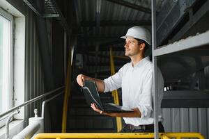 Smiling and happy employee. Industrial worker indoors in factory. Young technician with white hard hat. photo