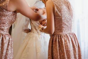 Bridesmaid helping slender bride lacing her wedding white dress, buttoning on delicate lace pattern with fluffy skirt on waist. Morning bridal preparation details newlyweds. Wedding day moments, wear. photo