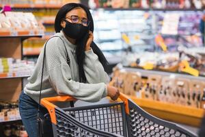 africano mujer vistiendo desechable médico máscara compras en supermercado durante coronavirus pandemia brote. epidemia hora foto