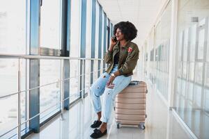 Portrait of happy young african american woman sitting on suitcase and talking with mobile phone at the station photo