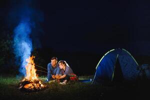 Pareja horneando salchichas en el fuego, y relajante por el hoguera en el bosque en el noche. romántico hoguera concepto. foto