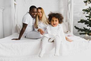 Portrait of happy multiracial young family lying on cozy white bed at home, smiling international mom and dad relaxing with little biracial girl child posing for picture in bedroom photo