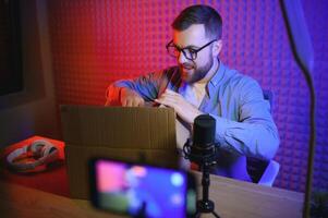 Panoramic view of young adult vlogger creating internet content, holding carton box in hands, unpacking received pack and recording media online video on broadcast equipment photo