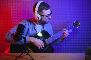 un joven elegante chico con un barba vistiendo casual o música profesor jugando guitarra en frente de teléfono inteligente cámara. en línea guitarra formación o vocal lección foto