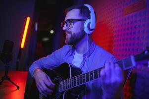 un joven elegante chico con un barba vistiendo casual o música profesor jugando guitarra en frente de teléfono inteligente cámara. en línea guitarra formación o vocal lección foto