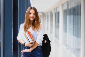 Positivity beautiful girl smiling at camera, standing on corridor with notes as backpack, going to lesson. Happy brunette female student studying in luxury university. photo