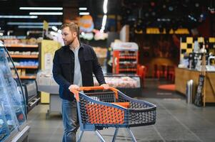 cliente en supermercado. hombre haciendo tienda de comestibles compras en pie con carro elegir comida producto adentro. chico comprando comestibles en comida almacenar. selectivo enfocar, Copiar espacio. foto