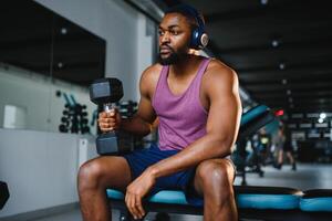 joven africano americano hombre sentado y levantamiento un pesa cerca a el estante a gimnasia. masculino peso formación persona haciendo un bíceps rizo en aptitud centro. foto