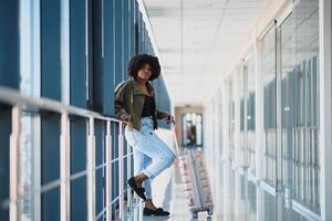 Young african american female passanger in casual clothes is in airport with baggage. photo