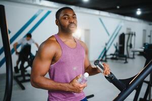 young african american sportsman drinking water and looking away in gym. photo