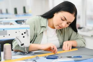 joven modista mujer cose ropa en trabajando mesa. sonriente costurera y su mano cerca arriba en taller. foto