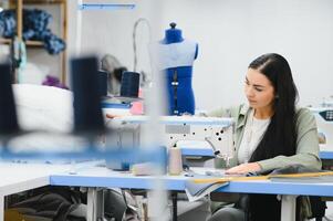 Cheerful young seamstress sits at the table with working things, in atelier photo