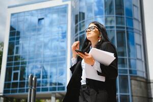 Young girl office worker in eyeglasses, dark jacket and blue shirt stays on city street near office building and holds her documents. photo