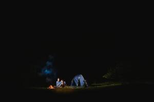 Camping night in mountains. Tourist couple sitting in front of illuminated tent lit by burning campfire. Tourism and outdoor activity concept. photo