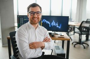 Man trader in formalwear sitting at desk in frot of monitors with charts and data at office browsing laptop checking documents analyzing stocks price changes concentrated. photo