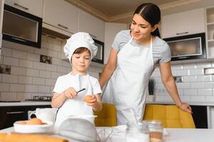 contento madre y su pequeño hijo sacudida crudo huevos en cuenco antes de haciendo masa para hecho en casa Pastelería en el cocina foto