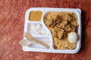 Indian Chicken Biryani packed in recycled box with raita and gravy and wooden spoon for home delivery. Top view photo