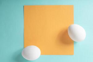 dos blanco huevos con amarillo tarjeta en un azul antecedentes. parte superior vista, plano poner, mínimo Pascua de Resurrección concepto foto