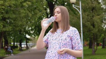 un' incinta donna nel un' estate vestito con un' floreale Stampa sta nel il parco e bevande puro primavera acqua arricchito con tracciare elementi. video