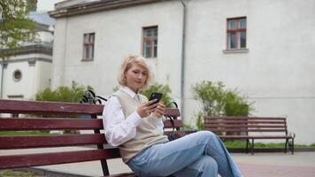 Jeune blond affaires femme dans une beige gilet et blanc chemisier est assis sur une banc dans le milieu de le ville et balayages dans une cellule téléphone avec café sur main dans main video