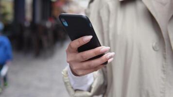 Young business woman in a beige trench coat and white blouse running in a cell phone standing on the street. Close-up view of the hand video