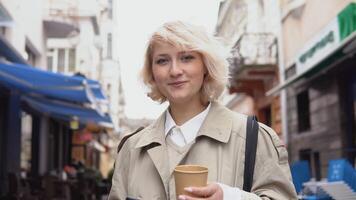 Jeune affaires femme dans une beige tranchée manteau et blanc chemisier avec une tasse de café et une cellule téléphone dans sa mains regards à le caméra tandis que permanent sur le rue. portrait de une souriant Jeune femme video