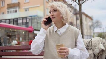 giovane bionda attività commerciale donna nel un' beige veste e bianca camicetta si siede su un' panchina nel il mezzo di il città e parla su un' cellula Telefono in linea con un' porta via caffè nel mano video