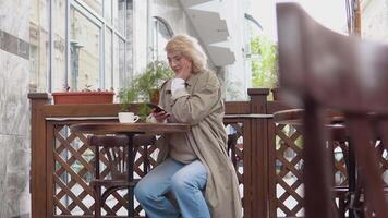 glad ung blond kvinna i tillfällig kläder svep i de telefon Sammanträde på en tabell med en kopp av kaffe på de terrass video