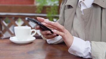 Woman swipes in the phone sitting at a table with a cup of coffee on the terrace. Close-up view of hands with cell phone. White porcelain cup on a background video