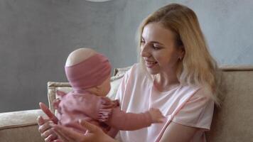 bebis flicka med blå ögon i en söt rosa kostym spelar med mamma på henne knä. mor och bebis i rosa kläder sitta motsatt varje Övrig på en mjuk ljus soffa video