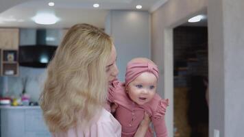 mãe balanços bebê dentro dela braços em a fundo do uma moderno cozinha definir. bebê menina com azul olhos dentro uma fofa Rosa terno sorrisos e parece às a Câmera video