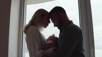 el madre y padre estar en frente de el ventana frente a cada otro y abrazo mientras el niño duerme Entre ellos en el de la madre brazos. paternidad y maternidad. tradicional familia video