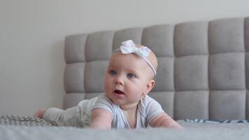 baby meisje in grijs kleren met een schattig boog Aan haar hoofd aan het liegen Aan de bed en op zoek Bij de camera. baby zorg. kind ontwikkeling in de eerste jaar van leven video