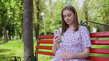 en gravid kvinna är Sammanträde i en sommar parkera och läsning information för gravid kvinnor på en smartphone. uppkopplad lektioner för förväntans föräldrar, mödrar och fäder video