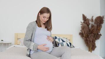 The pregnant woman is stroking her belly. Pregnant woman in home clothes sitting on a double bed. Dried flowers in the interior. Third trimester of pregnancy video