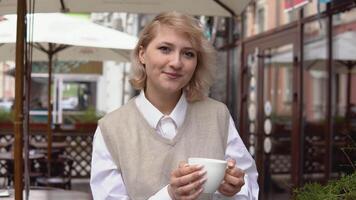 ung blond kvinna med elegant manikyr i en beige väst och vit blus innehav en vit porslin kopp med kaffe, stående på en sommar terrass i en gata Kafé. flicka ler, ser på de kamera video