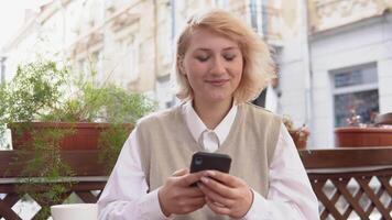 Jeune blond femme avec élégant manucure dans beige gilet et blanc chemisier travail en ligne dans cellule téléphone séance sur été terrasse dans une rue café video